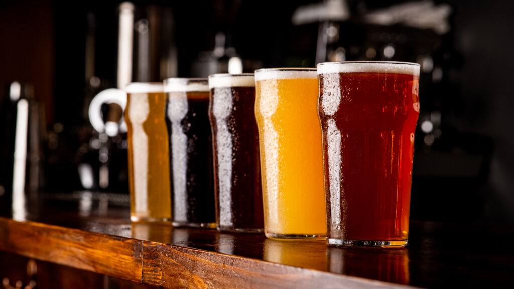 "A diverse array of beer glasses arranged in a gradient from light to dark, showcasing the spectrum of beer types. On the left, the lightest beer glows a pale yellow, transitioning through shades of amber and brown, to a rich, deep stout on the right. Each glass captures the unique clarity, effervescence, and head of its respective brew, illustrating the variety and complexity of beer flavors and styles."